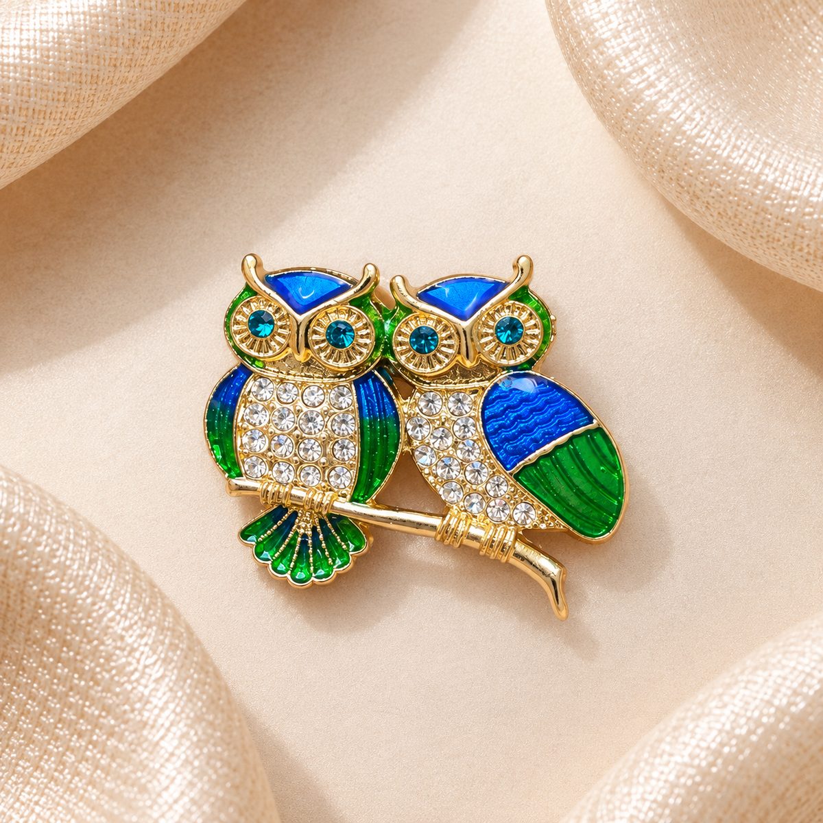 Broche deux chouettes dorées aux détails bleu et vert et aux strass, sur fond beige satiné