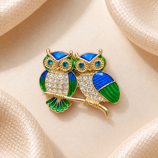 Broche deux chouettes dorées aux détails bleu et vert et aux strass, sur fond beige satiné