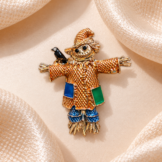 Broche épouvantail doré avec chapeau texturé, poches bleue et verte et petit corbeau noir sur fond champagne satiné