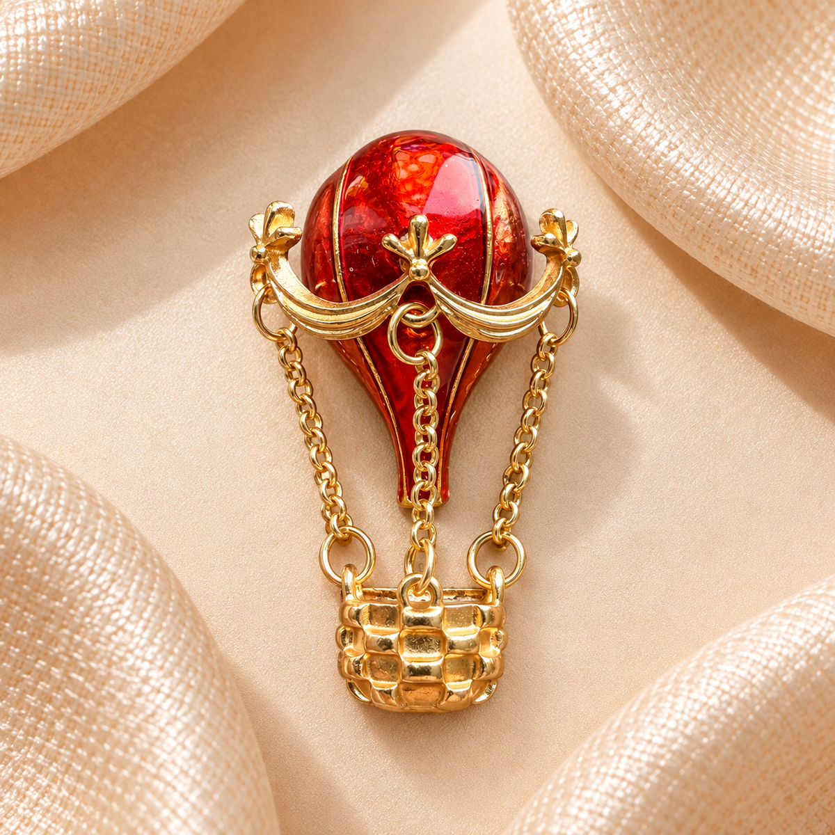 Broche montgolfière rouge rubis et dorée avec chaînes et panier sur fond satin champagne-beige